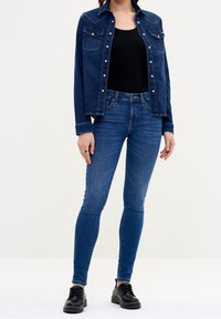 Chaqueta de mezclilla oscura con dos bolsillos en el pecho y cierres a presión, llevada sobre una camiseta sin mangas negra, combinada con jeans ajustados azul y zapatos negros.