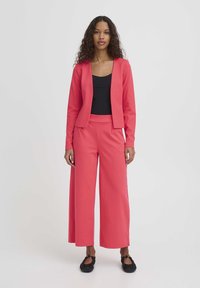 Ensemble deux pièces corail comprenant une veste cintrée sans fermetures et un pantalon à jambes larges, associé à un top noir ajusté et des chaussures plates noires.