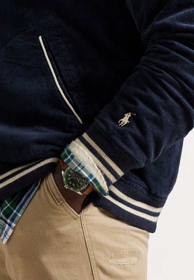 Navy corduroy jacket with cream stripes, left pocket, and embroidered logo. Cuff reveals plaid shirt. Silver watch with green face and brown strap.