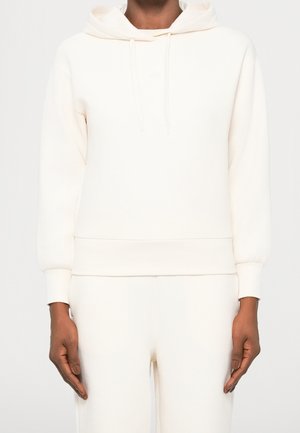 Personne portant un sweat à capuche uni de couleur crème et un pantalon assorti, debout devant un fond clair et neutre.