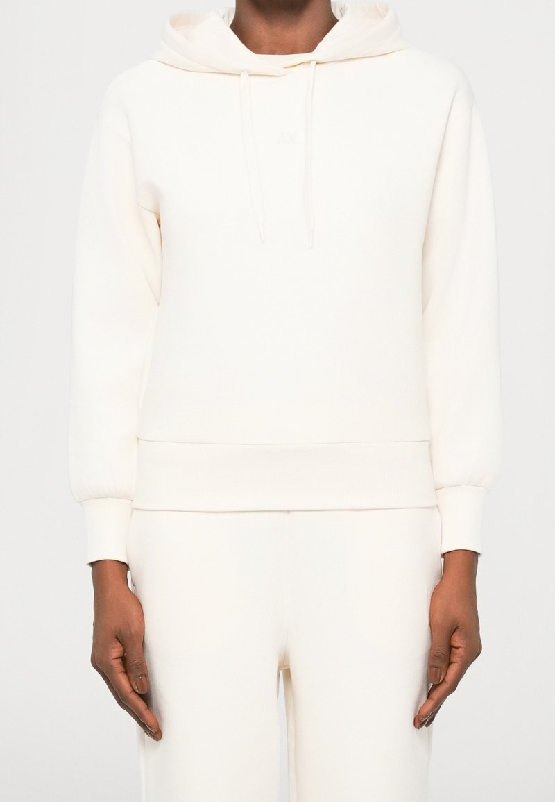 Personne portant un sweat à capuche uni de couleur crème et un pantalon assorti, debout devant un fond clair et neutre.