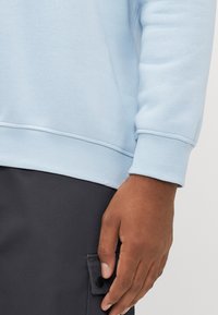 Brown-skinned hand resting near the pocket of dark pants, wearing a light blue long-sleeve sweatshirt with ribbed cuffs and hem.