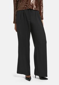 Pantalons noirs à jambes larges avec une texture lisse, ornés de plis à la taille ; assortis à des chaussures à talons aiguilles noires.