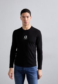 Black long-sleeve shirt made of cotton, featuring a front white logo design. Paired with blue fitted jeans, model poses against a neutral background.