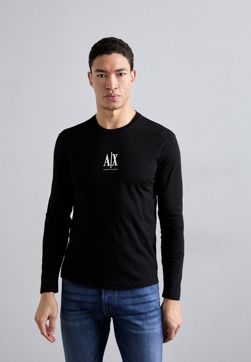 Black long-sleeve shirt made of cotton, featuring a front white logo design. Paired with blue fitted jeans, model poses against a neutral background.