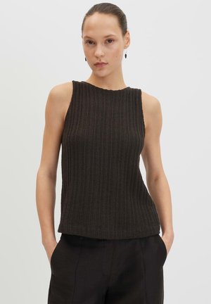 Femme aux cheveux attachés portant un haut noir en maille côtelée sans manches et un pantalon noir, debout les mains dans les poches sur fond blanc.