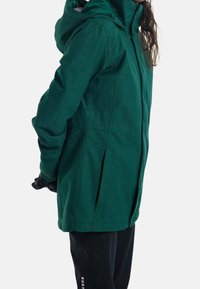Grüne Kapuzenjacke aus strukturiertem Stoff, mit Reißverschluss vorne und seitlichen Falten. Seitlich betrachtet, kombiniert mit schwarzen Hosen und Handschuhen.