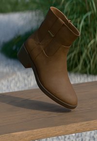 Καφέ δερμάτινη μπότα έως τον αστράγαλο με λείο υφή, στρογγυλή μύτη, μικρό τετράγωνο τακούνι και πλαϊκό φερμουάρ. Εμφανίζεται σε μια ξύλινη επιφάνεια.