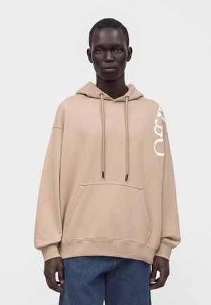 Persona que lleva una sudadera beige de gran tamaño con bolsillo frontal y cordones, combinada con vaqueros azules, de pie frente a un fondo liso.