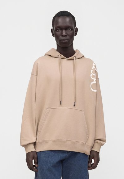 Personne portant un sweat à capuche beige oversized avec poche avant et cordons, associé à un jean bleu, debout devant un fond uni.