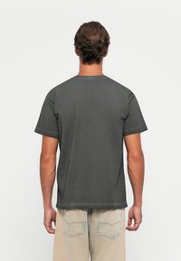 Homme aux cheveux bruns ondulés portant un t-shirt à manches courtes gris foncé uni et un jean beige clair, vu de dos contre un fond blanc.
