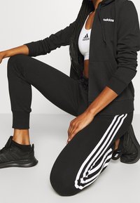 Survêtement noir avec une veste à capuche zippée et un pantalon ajusté arborant des rayures blanches sur les côtés. Accompagné de chaussures de sport noires.