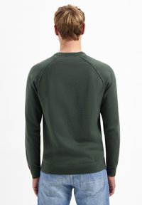 Donkergroene trui met een ronde hals, ribgebreide manchetten en onderrand. Kenmerkt zich door een gladde breistekstuur en raglanmouwen, gedragen met lichtblauwe jeans.