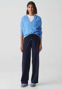 Pull moelleux bleu clair à col en V, associé à un pantalon large navy et des chaussures bleues à enfiler. Design minimaliste, texture douce.