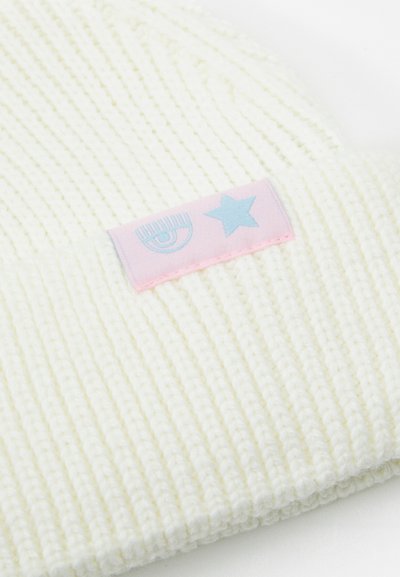 Bonnet en tricot couleur crème clair avec une texture côtelée. Présente une étiquette rose avec des accents graphiques d'étoile bleu clair et d'œil.