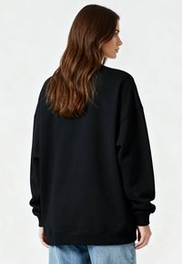 Femme aux longs cheveux bruns portant un sweat-shirt noir surdimensionné et un jean bleu clair, de profil sur un fond uni.