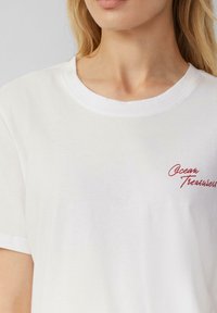 T-shirt en coton blanc avec un col rond. Présente un texte en cursive rouge "Ocean Treasures" sur le côté gauche. Manches courtes, texture douce.