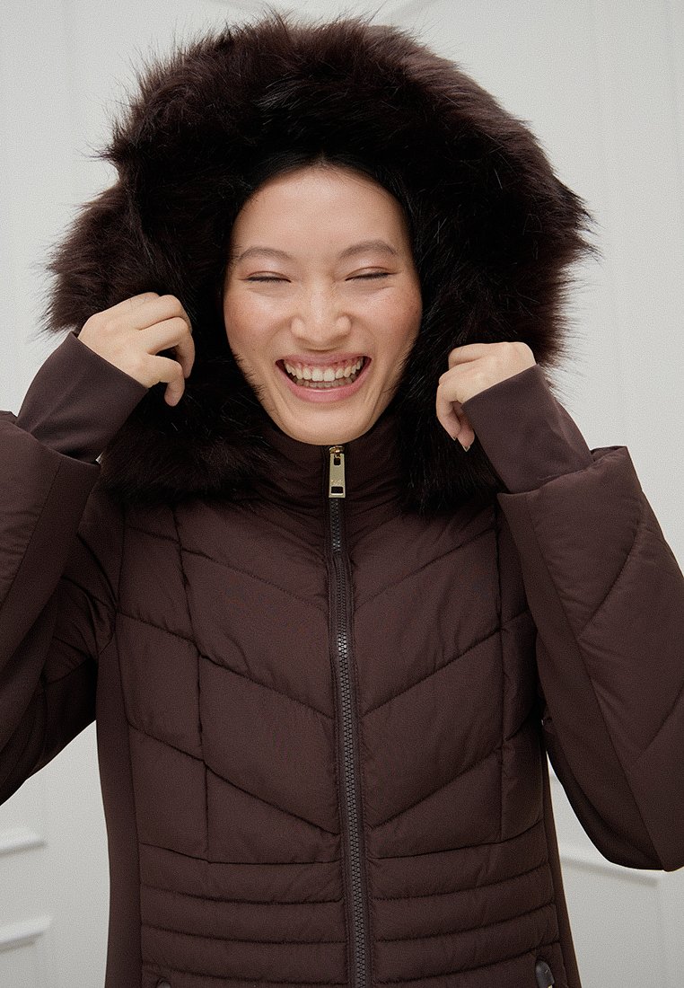 Glijdende vrouw in een donkerbruine gewatteerde winterjas met een met bont gevoerde capuchon, die de randen van de capuchon met beide handen vasthoudt.