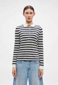 Tricou polo cu mâneci lungi, dungi în bleumarin și crem, cu plastron cu nasturi, guler moale și manșete refiletate. Combinație cu blugi deschis la culoare, nuanță de albastru deschis.