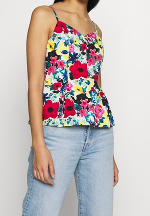 Kvinde iført en ærmeløs, farverig blomstret top med tynde stropper og lyseblå jeans, stående mod en ensfarvet baggrund.