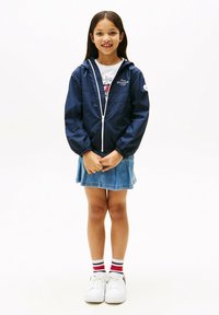 Coupe-vent marine avec une fermeture éclair à l'avant et des poignets élastiques, porté sur une chemise blanche, associé à une jupe en jean et des baskets blanches.