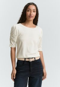 Romantische, korte mouwen top van zachte stof, met geplooide details op de schouders. Gecombineerd met donkere jeans en een lichte riem.