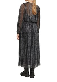 Schwarzes langes Kleid mit metallischem Glanz, durchsichtigen langen Ärmeln, gerafftem Taillenbereich und einem fließenden Rock. Kombiniert mit schwarzen klobigen Stiefeln.