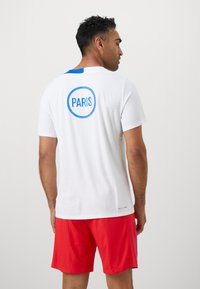 Hvid sportskjorte med blåt accent og "PARIS" trykt i en blå cirkel på ryggen, parret med røde teksturerede træningsshorts.