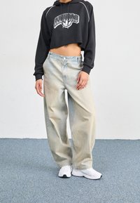 Czarna krótka bluza z kapturem z białym logo, jasnoniebieskie szerokie spodnie jeansowe oraz białe buty sportowe na niebieskim dywanie.