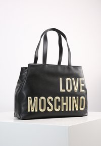 Černá kožená taška přes rameno s vyraženým nápisem "LOVE MOSCHINO" v zlaté barvě. Pevné dvojité úchyty a měkký, strukturovaný tvar.