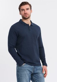 Man med kort brunt hår som bär en marinblå långärmad polo och blå jeans, står med en hand i fickan.