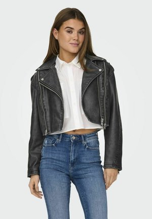 Veste en cuir noire courte de motard avec quincaillerie argentée, zips asymétriques et détails de couture, portée sur une chemise blanche et un jean bleu.