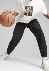 Bílý tričko s grafikou kočky držící basketbal, v kombinaci s černými jogger kalhotami a šedými sportovními botami s zelenými akcenty.