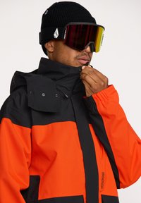 Veste de ski orange et noire avec un col haut, fermeture zippée et tissu texturé. Présente un logo sur la manche et une coupe ajustée.