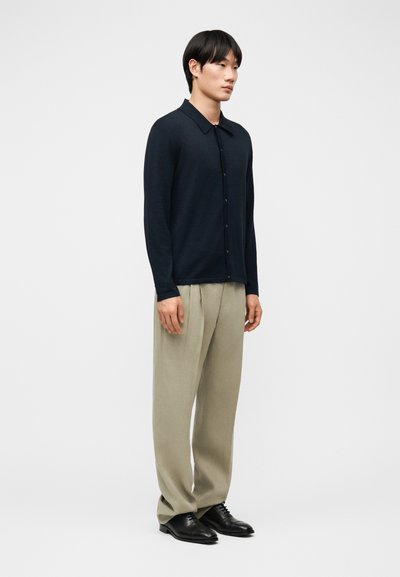 Άνδρας που στέκεται φορώντας μαύρη ζακέτα με κουμπιά, μπεζ πλισέ παντελόνι και μαύρα παπούτσια επίσημα, μπροστά σε απλό ανοιχτόχρωμο φόντο.