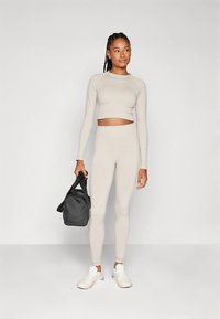 Lichtgrijs sportsetje met een cropped top met lange mouwen en hoge taille legging. Model houdt een zwarte sportschooltas vast en draagt witte sneakers.