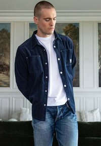 Camicia di velluto a coste color navy con due tasche frontali, chiusura a bottoni e colletto classico, indossata sopra una maglietta bianca e jeans blu.