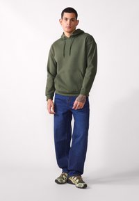 Dunkelgrüner Kapuzenpullover mit Kängurutasche, verstellbarem Kordelzug und gerippten Bündchen, kombiniert mit blauen weitem Jeans und mehrfarbigen Turnschuhen.