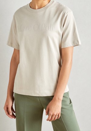 Une personne portant un T-shirt à manches courtes beige avec un texte en relief subtil et un pantalon vert olive devant un fond clair.