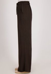 Losse donkerbruine broeken met elastische tailleband en zijzak, gedragen met beige espadrille flats, van opzij weergegeven op een effen achtergrond.