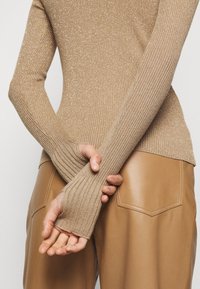 Personne portant un pull à manches longues côtelé beige et un pantalon en cuir marron, les mains croisées dans le dos, sur un fond neutre.