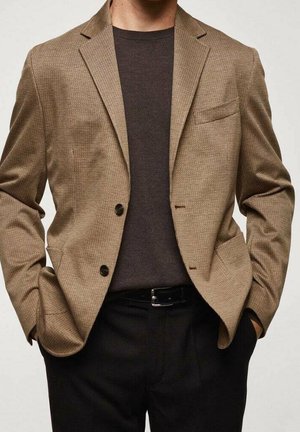 Mężczyzna w teksturowanej, wielbłądziej marynarce na ciemnobrązowym swetrze z okrągłym dekoltem, czarne spodnie, ręce w kieszeniach.