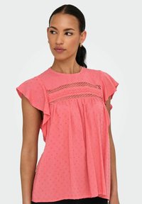 Blusa rosa a maniche corte con maniche arricciate, motivo a pois testurizzati e dettagli in pizzo decorativi sul petto. Tessuto leggero.