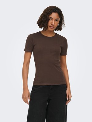 Jonge vrouw met kort krullend haar, gekleed in een aansluitend bruin topje met korte mouwen en losse zwarte broek tegen een effen lichte achtergrond.