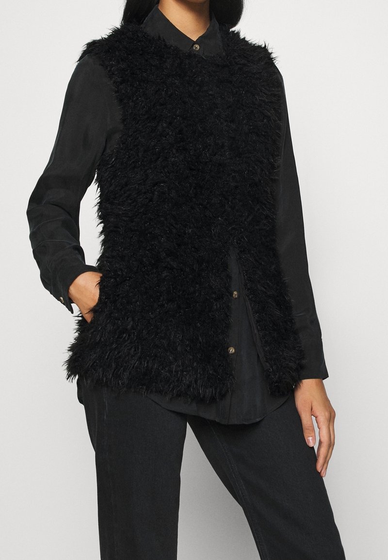 Black faux fur vest with a sleeveless design, featuring a high collar and soft texture, worn over a black button-up shirt and black pants.