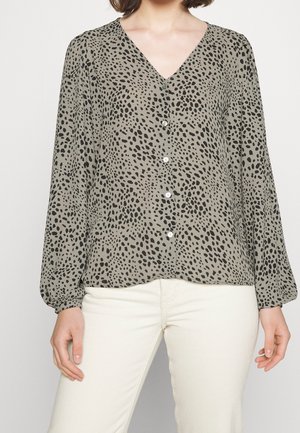 Woman wearing light olive green blouse with black irregular dot pattern and white buttons, paired with off-white pants.