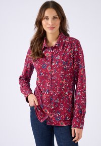 Donna con una camicia rossa a maniche lunghe a fiori con colletto, abbinata a jeans blu scuro, in piedi di fronte a uno sfondo neutro.