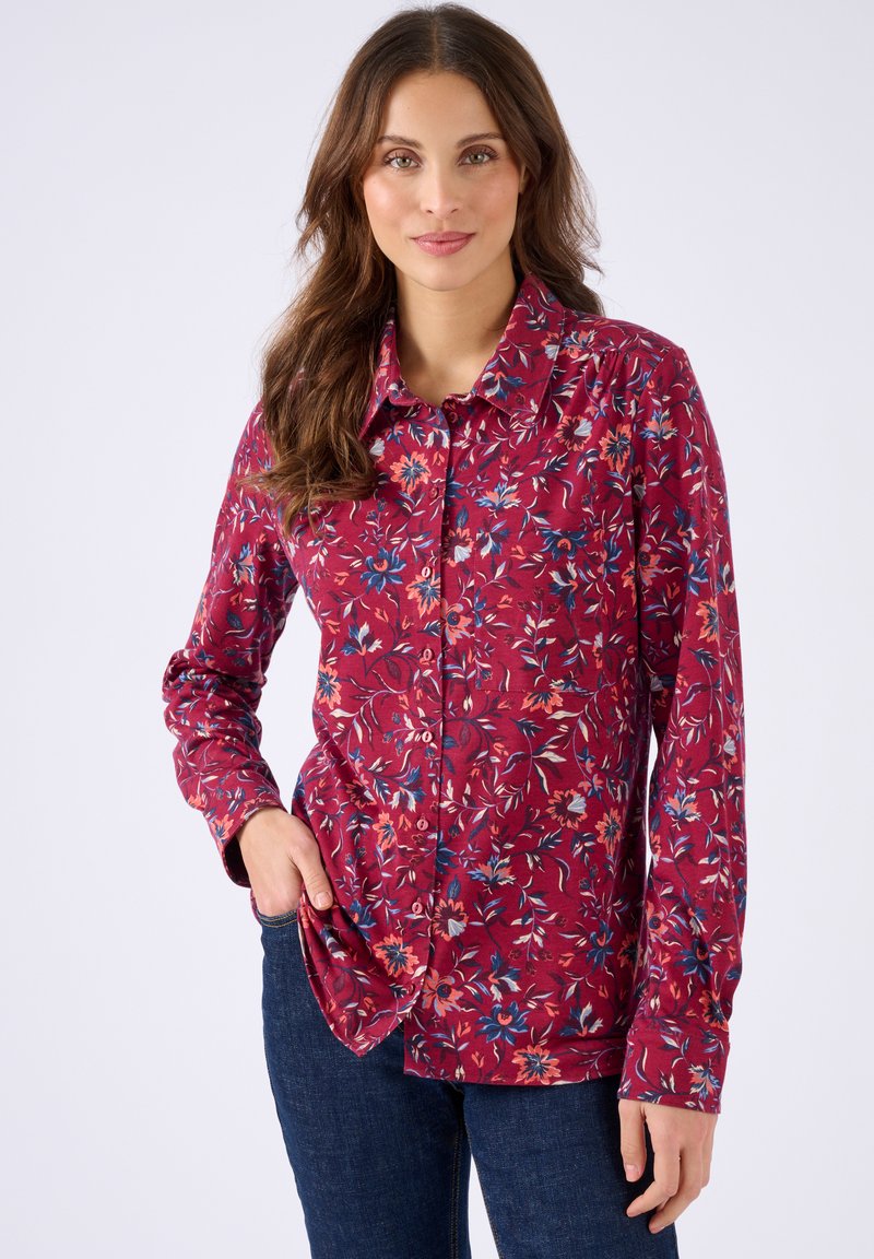 Donna con una camicia rossa a maniche lunghe a fiori con colletto, abbinata a jeans blu scuro, in piedi di fronte a uno sfondo neutro.