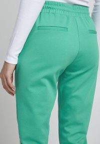 Rohelised spordipüksid, mis on valmistatud pehmest kangast, millel on sile tekstuur, koos kogutud vöökoht ja kahe tagataskuga.