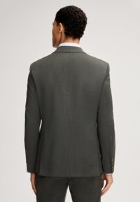 Mężczyzna w ciemnoszarym garniturze z białą koszulą, przedstawiony z tyłu na jednolitym jasnym tle.